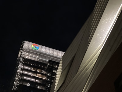 An urban scene featuring a tall building with modern architecture and illuminated windows. The brightly lit Google logo is prominently displayed on the top. The building is set against a dark night sky, and a nearby structure is visible on the right side with a textured surface.