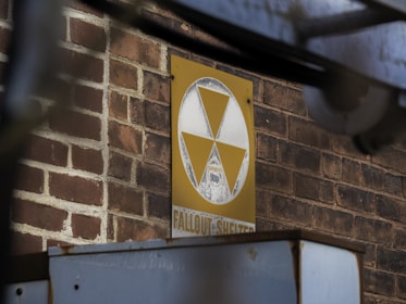 A vintage fallout shelter sign with a yellow and white triangle symbol is mounted on a brick wall. The sign appears weathered and aged, indicating historical significance. It reads 'Fallout Shelter' and displays a capacity number. Pipes or beams partially obstruct the view, giving an industrial or urban feel.