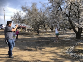 In a rural outdoor setting, a man is taking a photo with his camera of a person posing near a blooming tree with white flowers. There are other people, including children, nearby also enjoying the setting. Several parked cars are visible in the background. The ground is bare and appears dry, with some scattered fallen blossoms.