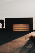 A modern garage door with a grid pattern is set against a minimalist building facade. The scene is captured during a time of day when long shadows stretch across the smooth concrete driveway, suggesting either dawn or dusk. Vegetation along the side adds a touch of nature to the stark architectural lines.