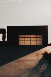 A modern garage door with a grid pattern is set against a minimalist building facade. The scene is captured during a time of day when long shadows stretch across the smooth concrete driveway, suggesting either dawn or dusk. Vegetation along the side adds a touch of nature to the stark architectural lines.
