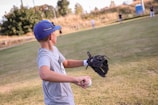 A young person wearing a blue cap and gray t-shirt is holding a baseball and a black glove on a grassy field. Several other individuals are in the background, also on the field likely participating in a baseball game. Trees and a blue structure are visible in the distance.