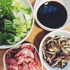 A wooden table contains four white bowls filled with various ingredients. One bowl has chopped green leafy vegetables, another contains dark sauce, a third holds sliced sausage, and the fourth has dried mushrooms.