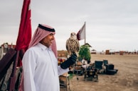 A man wearing a traditional white thobe and a red and white checkered keffiyeh is standing outdoors, holding a falcon on his gloved hand. Two flags are visible in the background along with a sandy landscape and some structures and greenery.