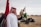A man wearing a traditional white thobe and a red and white checkered keffiyeh is standing outdoors, holding a falcon on his gloved hand. Two flags are visible in the background along with a sandy landscape and some structures and greenery.