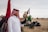 A man wearing a traditional white thobe and a red and white checkered keffiyeh is standing outdoors, holding a falcon on his gloved hand. Two flags are visible in the background along with a sandy landscape and some structures and greenery.