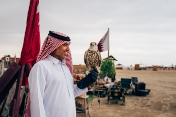 A man wearing a traditional white thobe and a red and white checkered keffiyeh is standing outdoors, holding a falcon on his gloved hand. Two flags are visible in the background along with a sandy landscape and some structures and greenery.