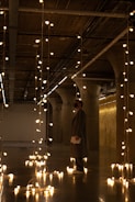 A person stands in an industrial-style space lit by numerous hanging Edison light bulbs, which create a warm, inviting atmosphere. The concrete columns and exposed ceiling fixtures add a modern, urban feel, while reflections from the light bulbs shine on the polished floor.