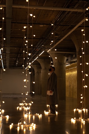 A person stands in an industrial-style space lit by numerous hanging Edison light bulbs, which create a warm, inviting atmosphere. The concrete columns and exposed ceiling fixtures add a modern, urban feel, while reflections from the light bulbs shine on the polished floor.