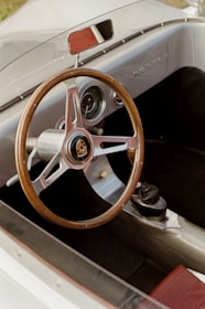 A vintage car interior is shown with a wooden steering wheel bearing the Porsche logo in the center. The dashboard features analog gauges, and a rear-view mirror is visible above the steering wheel. The interior comprises a combination of metallic and leather materials, with a red leather section visible near the seat.