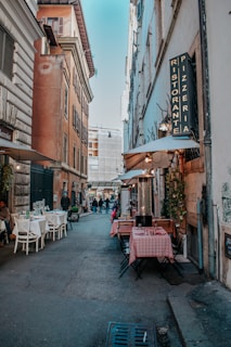 A narrow European street lined with outdoor tables from a restaurant. The tables have red and white checkered tablecloths. The street is bordered by tall, rustic buildings with old-style architecture. A sign for a 'Pizzeria Ristorante' hangs on the right side, and there are a few people strolling or sitting along the street. Strings of lights are visible, contributing to a cozy, welcoming atmosphere.