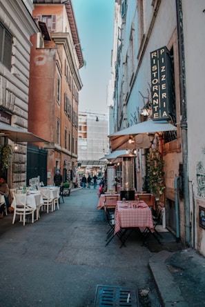 A narrow European street lined with outdoor tables from a restaurant. The tables have red and white checkered tablecloths. The street is bordered by tall, rustic buildings with old-style architecture. A sign for a 'Pizzeria Ristorante' hangs on the right side, and there are a few people strolling or sitting along the street. Strings of lights are visible, contributing to a cozy, welcoming atmosphere.