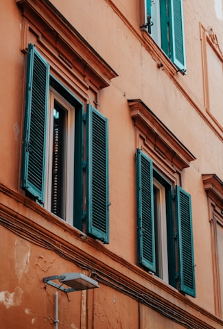 A building facade features textured orange walls with rustic appeal and multiple teal window shutters set against rectangular windows. The paint shows signs of weathering, adding character to the structure. Exposed cables run across the wall, and a simple wall-mounted lamp adds a functional element.