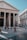 A grand ancient building with massive columns and Latin inscriptions on the facade. People are casually walking and standing in the open square in front of the structure, surrounded by cobblestone paving. The sky is clear and blue, creating a serene atmosphere.