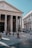 A grand ancient building with massive columns and Latin inscriptions on the facade. People are casually walking and standing in the open square in front of the structure, surrounded by cobblestone paving. The sky is clear and blue, creating a serene atmosphere.