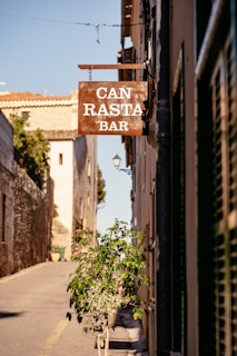 A street scene with a rustic sign for 'Can Rasta Bar' hanging above the sidewalk. The narrow street is lined with stone buildings and a small potted plant is visible. The atmosphere suggests a quaint, Mediterranean village with warm sunlight casting shadows on the ground.