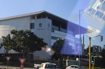 A modern building with the name Eduvos on its facade, surrounded by greenery and trees. There are road signs and traffic lights in the foreground. The reflection of a bus interior is visible on the right side.