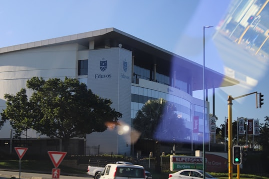 A modern building with the name Eduvos on its facade, surrounded by greenery and trees. There are road signs and traffic lights in the foreground. The reflection of a bus interior is visible on the right side.