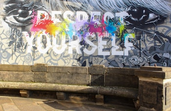 A vibrant wall mural features the large, bold text 'RESPECT YOURSELF' with splashes of colorful paint in red, yellow, blue, green, and purple across the words. Surrounding the text are black and white illustrations of eyes and cartoon-style characters, set against a detailed, patterned background. In front of the mural is an old, weathered stone bench and pavement.