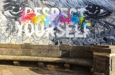 A vibrant wall mural features the large, bold text 'RESPECT YOURSELF' with splashes of colorful paint in red, yellow, blue, green, and purple across the words. Surrounding the text are black and white illustrations of eyes and cartoon-style characters, set against a detailed, patterned background. In front of the mural is an old, weathered stone bench and pavement.