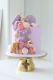 A pastel purple cake is elegantly decorated with various candies and chocolates, including chocolate bars, cake pops, pretzels, and popsicles. There are colorful sprinkles and edible gold leaf elements. The cake is set on a shiny, gold cake stand.