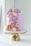 A pastel purple cake is elegantly decorated with various candies and chocolates, including chocolate bars, cake pops, pretzels, and popsicles. There are colorful sprinkles and edible gold leaf elements. The cake is set on a shiny, gold cake stand.
