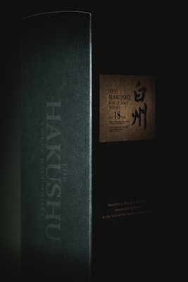 A dark and moody packaging of a single malt whisky with distinct branding. The box features Japanese characters alongside English text, indicating it is aged 18 years. The minimal lighting highlights the name 'The Hakushu' on the side and a textured label on the front.