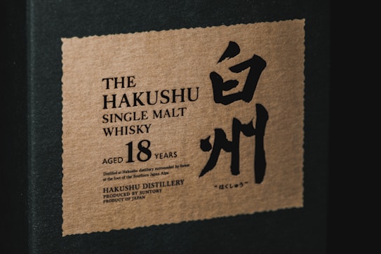A close-up of a whisky box featuring a label with the words 'The Hakushu Single Malt Whisky', aged 18 years. The design includes both English and Japanese characters, with a minimalist aesthetic on a textured background.