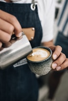 A barista wearing a denim apron is skillfully pouring milk into a cup of coffee to create latte art with a heart design. The focus is on the hands, the milk pitcher, and the ceramic cup, which has a textured, stone-like appearance.