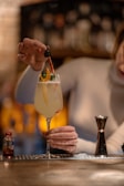 A person in a long sleeve shirt is holding a cocktail glass with a citrus garnish and using a dropper to add liquid to the drink. A small bottle and a jigger are on a bar counter, with a blurred background featuring bottles on shelves.