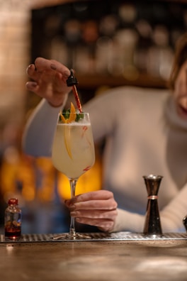 A person in a long sleeve shirt is holding a cocktail glass with a citrus garnish and using a dropper to add liquid to the drink. A small bottle and a jigger are on a bar counter, with a blurred background featuring bottles on shelves.