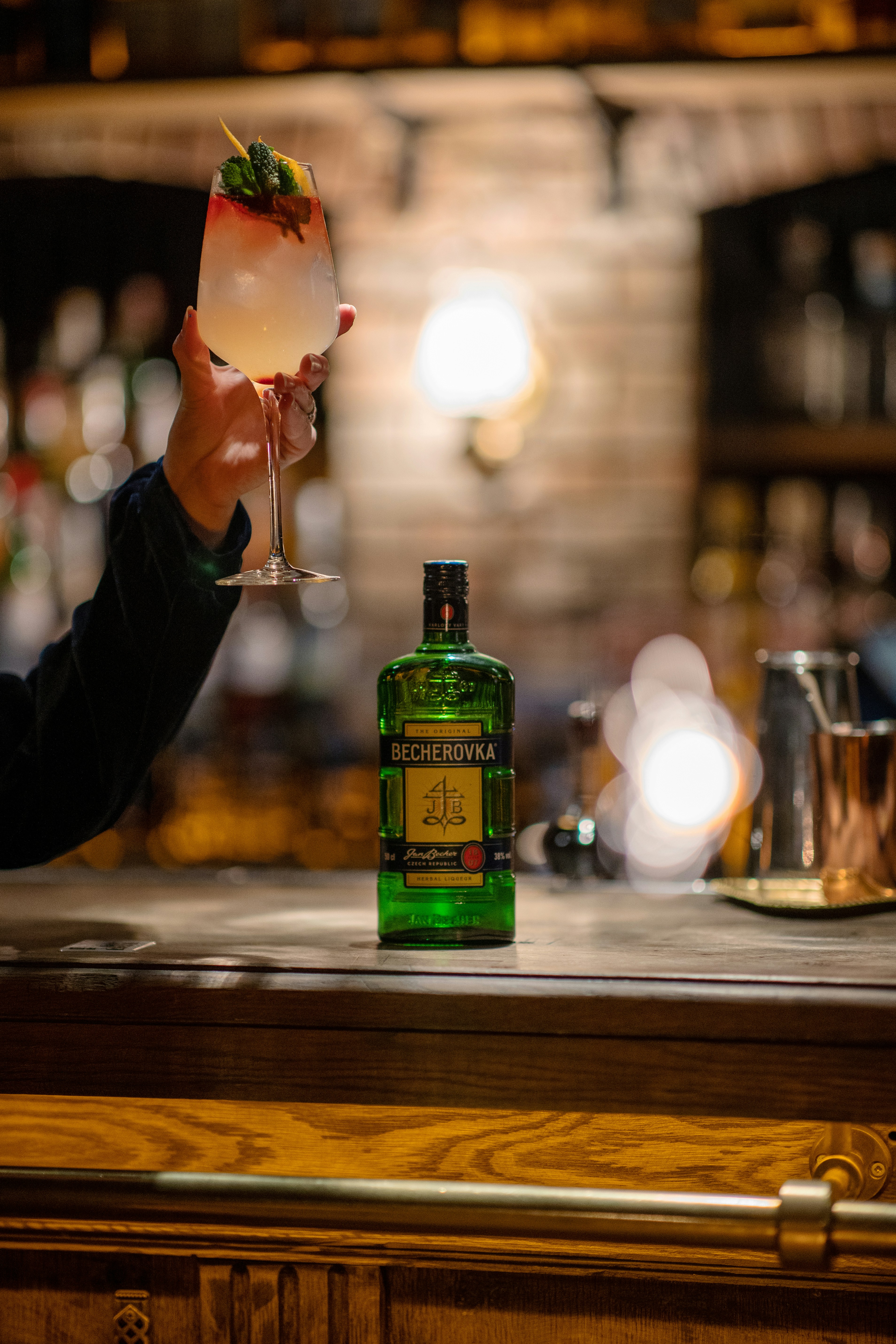 A hand is holding a cocktail glass with a layered white and red drink, garnished with mint leaves and a lemon twist. The setting appears to be a dimly lit bar with a rustic ambiance. A bottle of Becherovka is placed on the wooden bar counter, with some copper bar accessories visible in the background.
