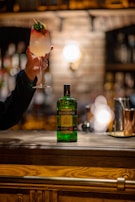 A hand is holding a cocktail glass with a layered white and red drink, garnished with mint leaves and a lemon twist. The setting appears to be a dimly lit bar with a rustic ambiance. A bottle of Becherovka is placed on the wooden bar counter, with some copper bar accessories visible in the background.