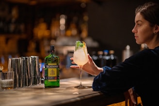A person sitting at a dimly lit bar holds a cocktail in their hand. The bar counter has several stainless steel shakers and a bottle labeled 'Becherovka' in the foreground. The person's focus appears to be on the drink, which is garnished with mint and lemon or lime.