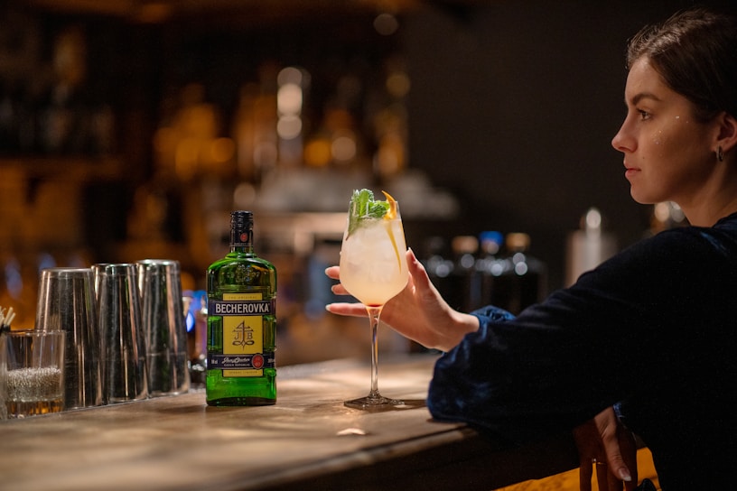 A person sitting at a dimly lit bar holds a cocktail in their hand. The bar counter has several stainless steel shakers and a bottle labeled 'Becherovka' in the foreground. The person's focus appears to be on the drink, which is garnished with mint and lemon or lime.
