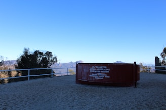 A large, reddish-brown telescope mirror display is positioned on a viewing platform overlooking a scenic landscape. The inscription reads '154'' Diameter Anglo-Australian Telescope Mirror, Grubb Parsons, Newcastle Upon Tyne, England'. In the background, there are mountains and trees under a clear blue sky.
