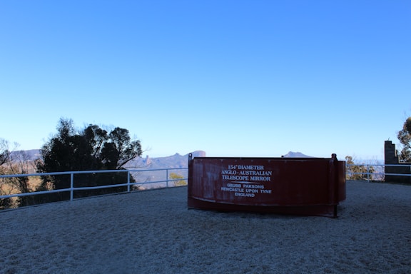 A large, reddish-brown telescope mirror display is positioned on a viewing platform overlooking a scenic landscape. The inscription reads '154'' Diameter Anglo-Australian Telescope Mirror, Grubb Parsons, Newcastle Upon Tyne, England'. In the background, there are mountains and trees under a clear blue sky.