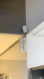 A modern indoor space with a sleek design, featuring a sign that reads 'Return' with Korean characters beneath it. The environment appears clean and organized, with a metallic and white color scheme.