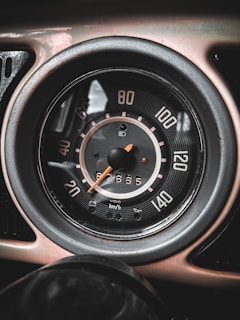 The image features a close-up view of a vintage car speedometer with a black face, white numbers, and an orange needle. It displays speeds in kilometers per hour ranging from 0 to 140, with the needle pointing slightly below 60. The odometer shows a reading of 62665. The entire gauge is encased in a metallic rim.