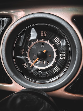 The image features a close-up view of a vintage car speedometer with a black face, white numbers, and an orange needle. It displays speeds in kilometers per hour ranging from 0 to 140, with the needle pointing slightly below 60. The odometer shows a reading of 62665. The entire gauge is encased in a metallic rim.