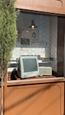 A retro computer sits in a window display with a hexagonal tile pattern wall. Above the computer, a vintage-style lamp hangs, and various posters with text and images are pinned to the wall. A small tree is partially visible on the left side of the image.