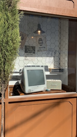 A retro computer sits in a window display with a hexagonal tile pattern wall. Above the computer, a vintage-style lamp hangs, and various posters with text and images are pinned to the wall. A small tree is partially visible on the left side of the image.