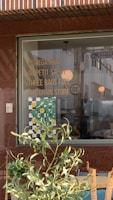 A storefront window displays a list of names or phrases like MOHEGANSUN, AU PETIT SALUT, THREE BAGS FULL, and THE GRAIN STORE in bold yellow lettering. Below these, there is a decorative element featuring a checkered pattern with green leaves and smiling yellow faces. In front of the window, green leafy plants partially obscure the view.