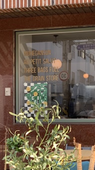 A storefront window displays a list of names or phrases like MOHEGANSUN, AU PETIT SALUT, THREE BAGS FULL, and THE GRAIN STORE in bold yellow lettering. Below these, there is a decorative element featuring a checkered pattern with green leaves and smiling yellow faces. In front of the window, green leafy plants partially obscure the view.