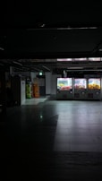 Dimly lit indoor area with a row of vending or claw machines on the right displaying brightly colored items. The floor is reflective, and there are stacks of colorful bottles or containers to the left. The space has a slightly industrial or commercial appearance.