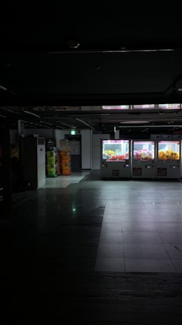 Dimly lit indoor area with a row of vending or claw machines on the right displaying brightly colored items. The floor is reflective, and there are stacks of colorful bottles or containers to the left. The space has a slightly industrial or commercial appearance.