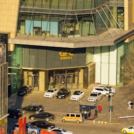 A modern glass building with the letters 'GFC' displayed above the entrance. Several cars are parked in front of the entrance, and a few people are walking nearby. The facade is made up of large windows and steel beams.