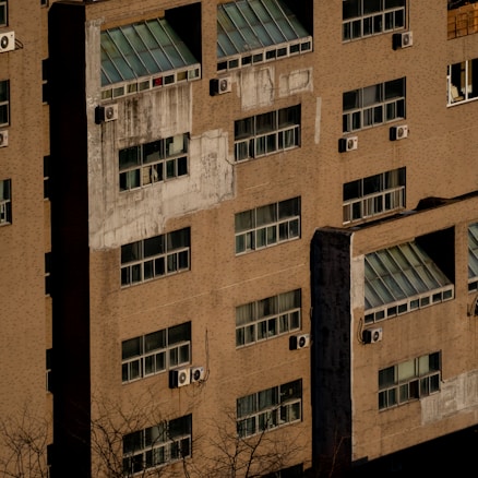 A close-up view of an old building exterior with multiple windows, some covered with grime and others displaying window air conditioning units. The structure's facade appears weathered and has visible patches of discoloration. Leafless tree branches are visible in the foreground.