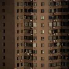 A high-rise apartment building with numerous windows, each adorned with either curtains or blinds. Several air conditioning units are affixed to the exterior of the building. The facade is uniformly colored, with variations in lighting due to the time of day or weather conditions.