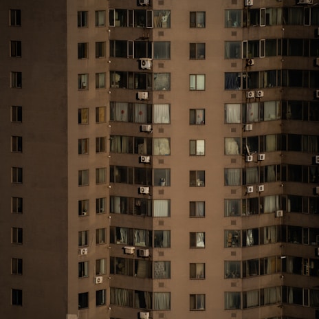 A high-rise apartment building with numerous windows, each adorned with either curtains or blinds. Several air conditioning units are affixed to the exterior of the building. The facade is uniformly colored, with variations in lighting due to the time of day or weather conditions.